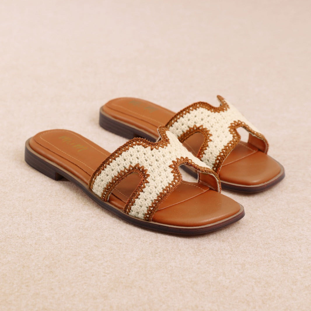 A pair of women's slide sandals with brown leather straps and a cream woven H-shaped detail, set against a neutral beige background.