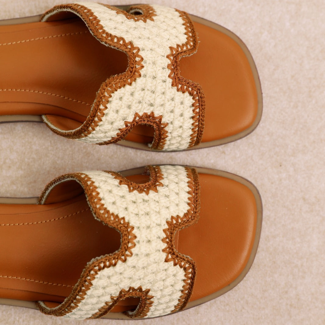 A pair of women's slide sandals with tan soles and straps featuring a cream and brown crocheted zigzag pattern, shown from a top-down view on a light beige textured background.